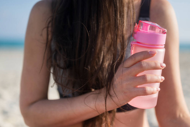 girl in sportswear with drinking water in a bottle on the beach near the sea - Zdjęcie, obraz