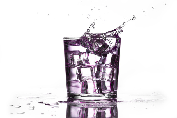 A crystal glass with ice and a purple liquid splashing with the fall of an ice cube, isolated on a white background and reflected on the surface. - Zdjęcie, obraz
