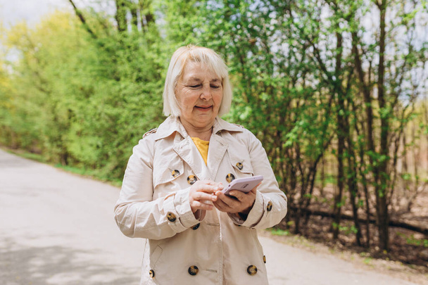 Mature blonde woman in casual clothes browsing smart phone while park of city. Caucasian blogger holding phone pondering thinking on idea for social publication outdoors - Foto, immagini