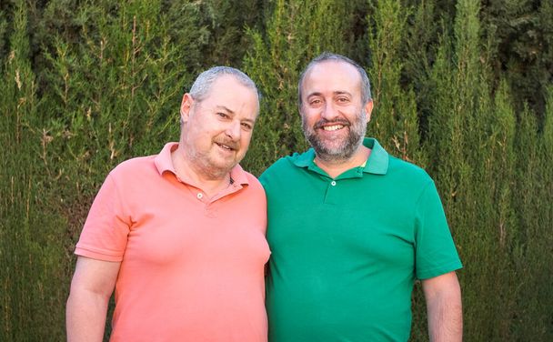Smiling bearded male friends in casual wear looking at camera while standing near green plants in rural area on summer day - 写真・画像