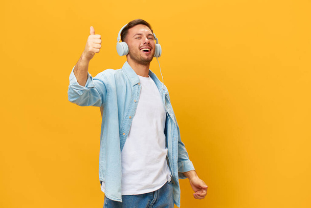 Dancing happy tanned handsome man in blue basic t-shirt headphones show thumb up gesture Like posing isolated on orange yellow studio background. Copy space Banner Mockup. Time to relax concept - Photo, Image