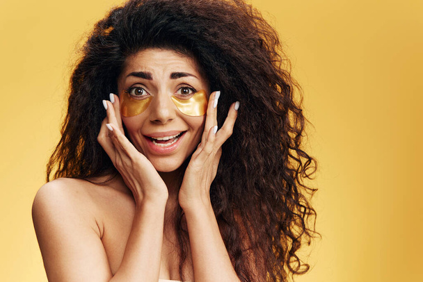 Verwirrt glücklich gebräunte hübsche Latin Lady berühren goldene Hydrogelpflaster unter den Augen, die isoliert vor gelbem Hintergrund posieren. Kosmetik Produktanzeige Natürliches Schönheitskonzept Studio Portrait Raum kopieren - Foto, Bild
