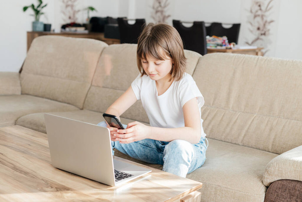 Mesafe öğrenme. Kız evde kanepede oturur ve dizüstü bilgisayar kullanarak ders çalışır ya da telefon eder ve saat dersleri alır. Çocuklar teknoloji kullanır.. - Fotoğraf, Görsel