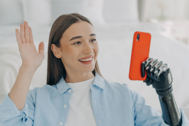 Young disabled woman has video call on phone. European girl is holding the mobile phone with bionic artificial arm and taking selfie. Happy caucasian woman with artificial limb at home. - Valokuva, kuva