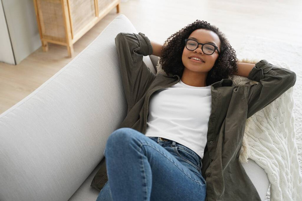 Relaxed biracial teen girl in glasses lying on comfortable couch, hands behind her head, relax on sunny lazy weekend at home. Serene young woman relaxing on cozy sofa in living room enjoy break. - Foto, imagen