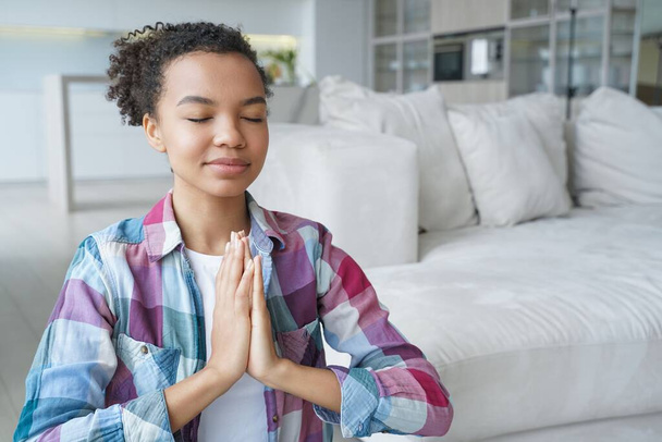 Spanish young woman is sitting on floor in lotus position with her eyes closed. Teenage afro girl is practicing yoga at home. Tranquility and meditation. Stress relief and mental health concept. - Foto, Imagem