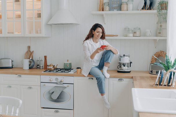 Caucasian young woman has message on cellphone. Happy girl with telephone sitting on worktop of table at kitchen and texting. Modern luxurious scandinavian interior. Lifestyle concept. - Photo, Image
