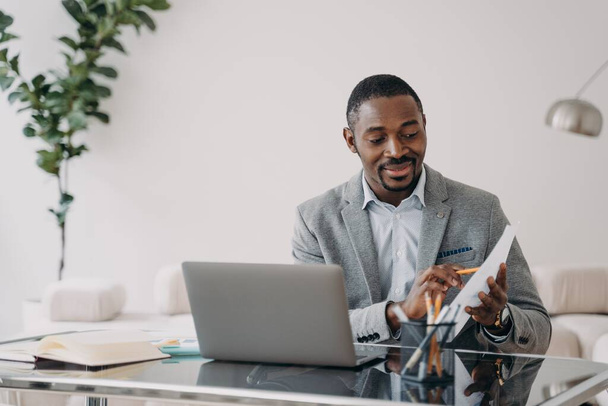 Businessman has paperwork. Happy man has video call on laptop and pointing to data papers with pencil. Freelancer is working online at home office. Remote african american worker. - Photo, Image