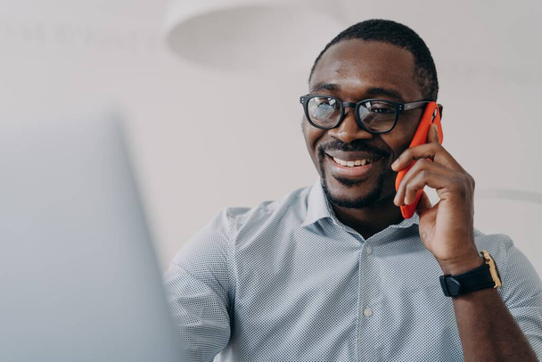 Successful african american businessperson in glasses hold mobile phone makes nice business call sitting in front of laptop. Black male friendly manager consulting a client, talking on smartphone. - Photo, image