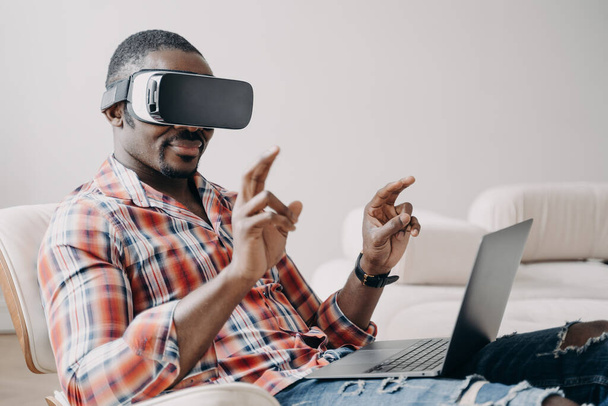 African american guy software delevoper wearing virtual reality glasses working in cyberspace at laptop. Modern black guy programmer freelancer developing programming VR game or application. - 写真・画像