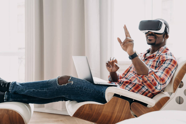 VR kulaklıklı Afro-Amerikan bir adam ev ofisinin vizyonundan zevk alıyor. İş adamının sanal toplantısı var. Rahat serbest çalışan biri sandalyede oturuyor ve proje üzerinde çalışıyor. Yüksek teknoloji dijital teknoloji. - Fotoğraf, Görsel