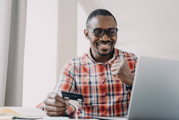 Afro guy gets profit. Happy african american man is buying or ordering online with pc. Businessman has transaction using credit card from home. Ecommerce and consumerism concept. Internet banking. - Photo, Image