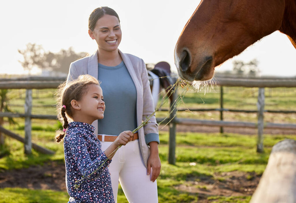 an adorable little girl feeding a horse on her farm while her mother looks on. - Photo, image