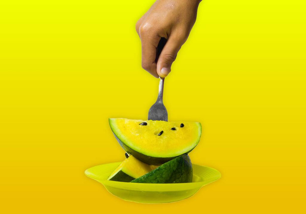 hand holding a fork stuck in a delicious and healthy yellow watermelon isolated on an orange yellow background. the benefits of watermelon help heart health and can nourish the body's skin - Fotó, kép