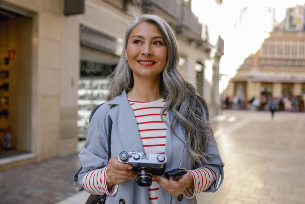 Good looking Asian woman with long grey hair in trench coat holding camera and looking away on street of old town - Fotoğraf, Görsel