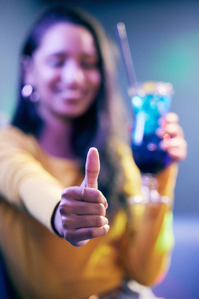 Μια νεαρή γυναίκα πίνει κοκτέιλ και δείχνει τους αντίχειρες της σε μια διασκεδαστική έξοδο.. - Φωτογραφία, εικόνα