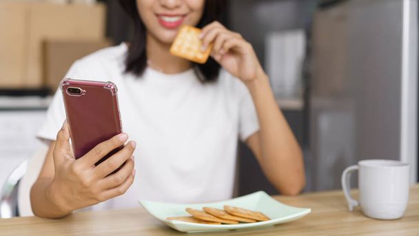 Kodikas elämäntapa käsite, Nuori nainen syö keksejä ja surffaa sosiaalisessa mediassa älypuhelimella kotona. - Valokuva, kuva