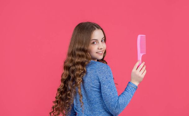positive teen girl with curly hair hold comb on pink background. - Фото, изображение