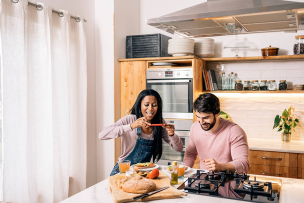 Cheerful black woman and her boyfriend having delicious breakfast in the kitchen at home and taking photo. - Fotó, kép