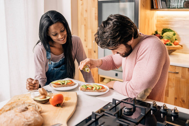 Cheerful black woman and her boyfriend having delicious mediterranean breakfast in the kitchen at home. - Foto, Imagen