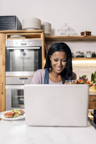 Happy black woman drinking juice in the kitchen at home and using computer. - Φωτογραφία, εικόνα