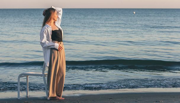 Красива жінка стоїть біля моря на заході сонця на тлі блакитних хвиль
. - Фото, зображення