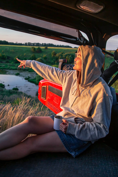 Mladá žena si užívá západ slunce, zatímco sedí v kufru SUV na venkově. - Fotografie, Obrázek