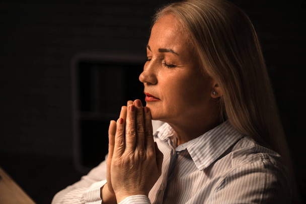 Religious mature woman praying in dark room - Zdjęcie, obraz