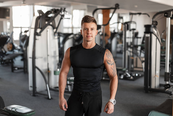Πορτρέτο ενός όμορφου μοντέρνου αθλητικού μοντέλου με χτένισμα και τέλειο μυϊκό σώμα σε μαύρο αθλητικό ένδυμα με μπλουζάκι μπροστά από το γυμναστήριο - Φωτογραφία, εικόνα