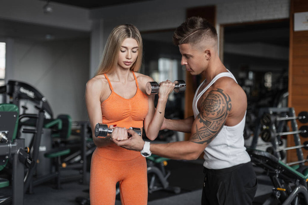 Athletic young beautiful couple in sportswear working out with metal dumbbells in the gym.  Beautiful girl trains with a fitness trainer. Handsome guy helps a girl in the gym - Photo, Image