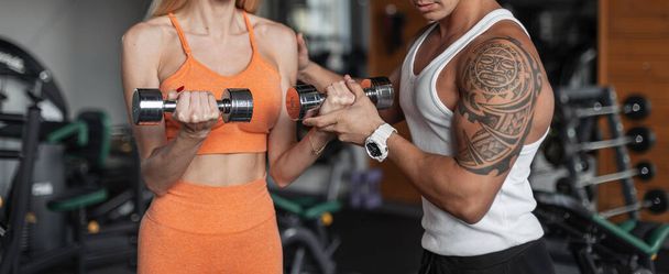 Women's hands holding metal dumbbells and pumping her biceps with a fitness trainer. Guy helps a girl work out at the gym - 写真・画像