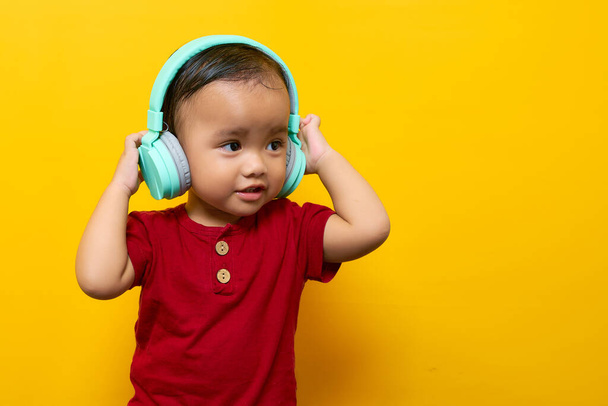Веселый молодой азиат, 2-летний мальчик в красной футболке, слушает любимую музыку с наушниками, изолированными на желтом фоне. День матери - это семейный образ жизни - Фото, изображение