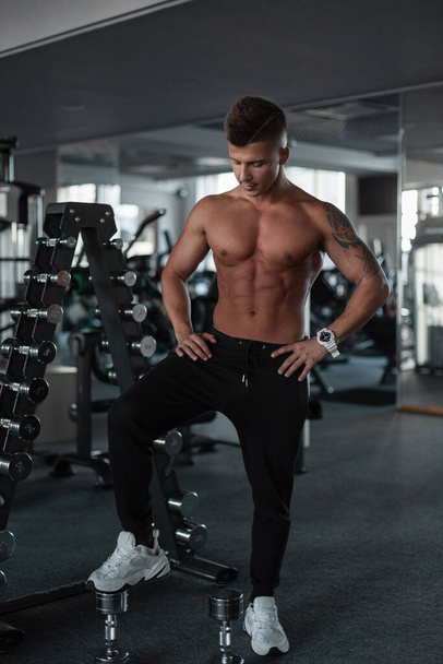 Sporty handsome strong male bodybuilder with a sexy muscular body with shorts, tights and sneakers posing and standing with his foot on dumbbells in the gym - Фото, зображення
