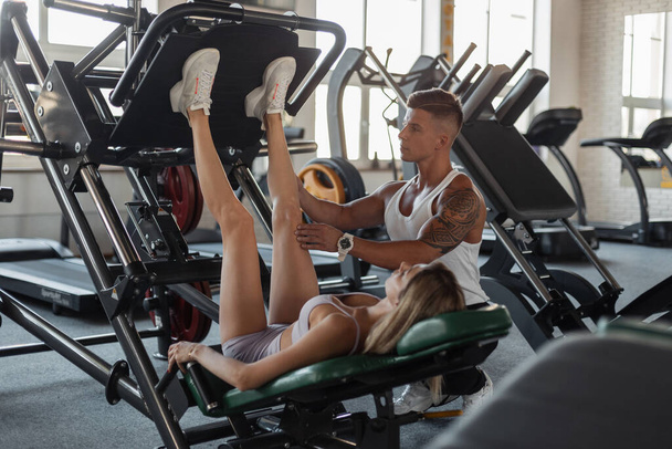 Erkek fitness antrenörü güzel sarışın bir kadını eğitir ve spor salonundaki bir makineye bacak kaslarını pompalar. - Fotoğraf, Görsel