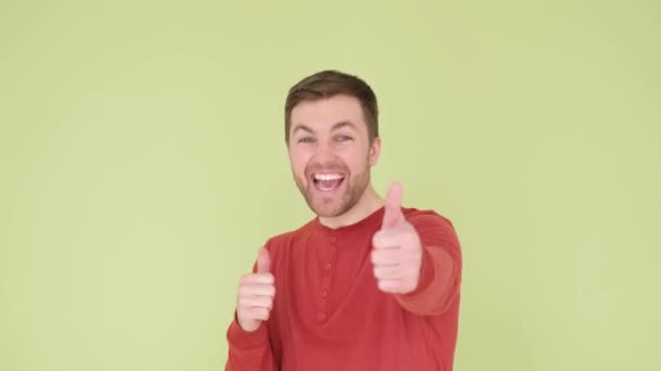 Handsome man with a beard in an orange sweater on a yellow-green background, shows a sign of success, makes a positive gesture. Smiling and happy guy thumb up. Cheerful facial expression - 映像、動画