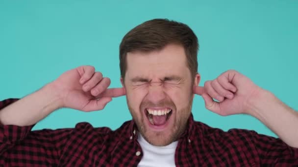 Angry man shouting and covering his ears with his hands isolated on a turquoise background in the studio. Close up of nervous man screaming loudly at camera, unhappy man. 4k video - Video, Çekim