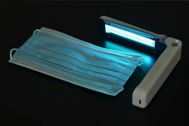 Portable quartz lamp with blue light disinfect medical mask on dark background from bacteria, virus, coronavirus, healthcare concept - Foto, Imagem