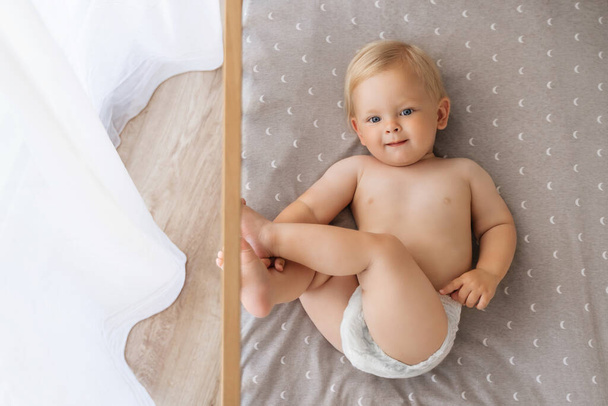 Upper view of sweet cute blue-eyed baby in diaper lying on his back in crib looking at camera, playing with foot putting legs up. Childcare concept. Happy carefree childhood. Copy space - Zdjęcie, obraz