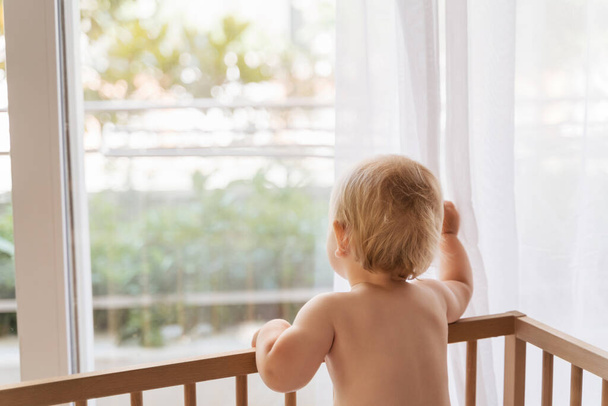 Πίσω όψη του λυπημένου μοναχικού μωρού που στέκεται στο κρεβάτι κοιτάζοντας μέσα από το παράθυρο περιμένοντας με ανυπομονησία την επιστροφή της μαμάς ή του μπαμπά του, που έμεινε μόνο στο σπίτι. Παιδικό τραύμα. Κακές οικογενειακές σχέσεις. Παιδική μέριμνα - Φωτογραφία, εικόνα