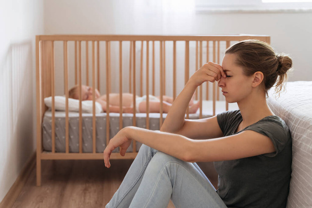 Side view of unhappy frustrated young mother sitting on floor in child bedroom while baby sleeping in bed, trying to calm down, suffering postnatal depression symptoms - Фото, изображение