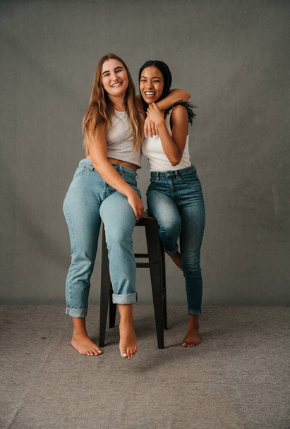 Two girl friends smiling and wearing jeans and white tops sharing a stool in the studio. High quality photo - Foto, Imagen