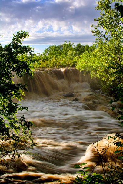 The vertical view of the rocky river waterfalls surrounded by trees under the cloudy sky - Fotó, kép