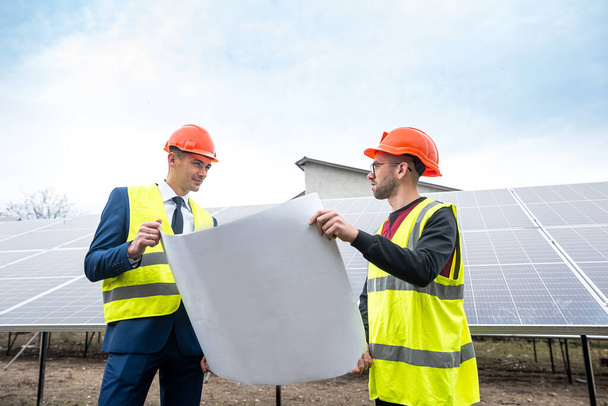 the boss in a helmet and vest stands with an employee in special clothes near the solar panels. Green electricity concept - Φωτογραφία, εικόνα