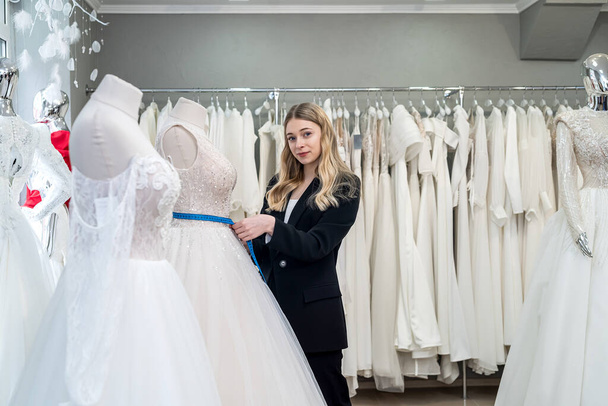 female tailor measuring elegant wedding dress on mannequin using tape measure in store - 写真・画像