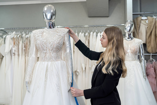 Charming woman saleswoman in stylish black suit measures wedding dress with measuring tape in boutique - Photo, Image