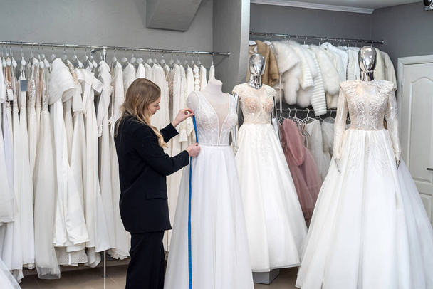Schneiderin arbeitet im Hochzeitssalon mit zentimetergroßem Maßband im weißen eleganten Kleid - Foto, Bild