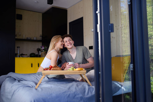 A young beautiful couple in love is sitting in bed and having healthy breakfast together. - Фото, изображение