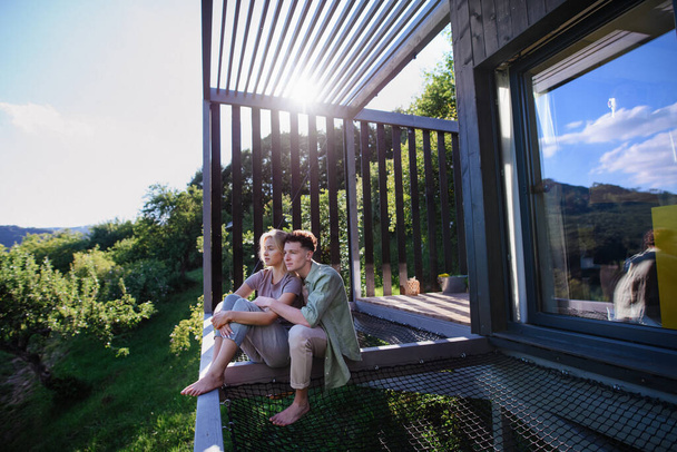A young couple sitting and cuddling in hammock terrace in their new home in tiny house in woods, sustainable living concept. - Foto, Imagen