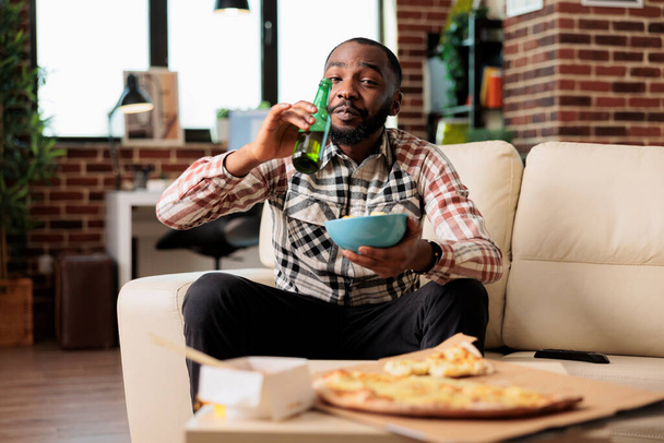 Happy guy drinking alcoholic beer from bottle and holding bowl of chips, watching film on television program. Enjoying beverage with takeaway delivery food and snacks, entertainment. - Photo, Image