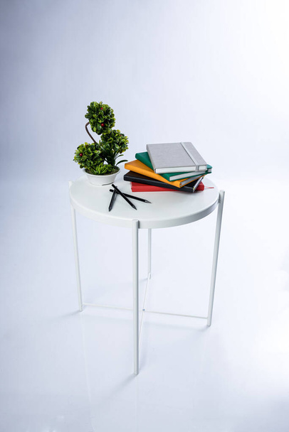 Stack of colourful notebooks and pens on a white table. A plant is next to them. Product photoshoot in a studio. High quality photo - Foto, Imagen